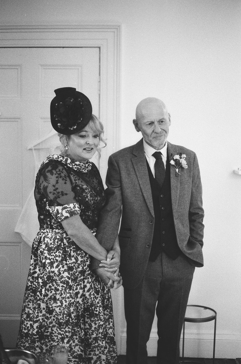 Black and white photo of a formal occasion showing a woman in a patterned dress and small hat standing beside man in suit.