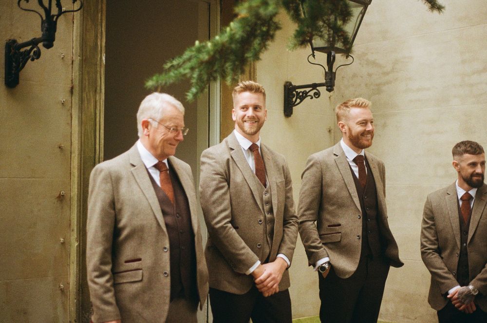 Groomsmen in matching gray suits standing together outside a stone building at a wedding.