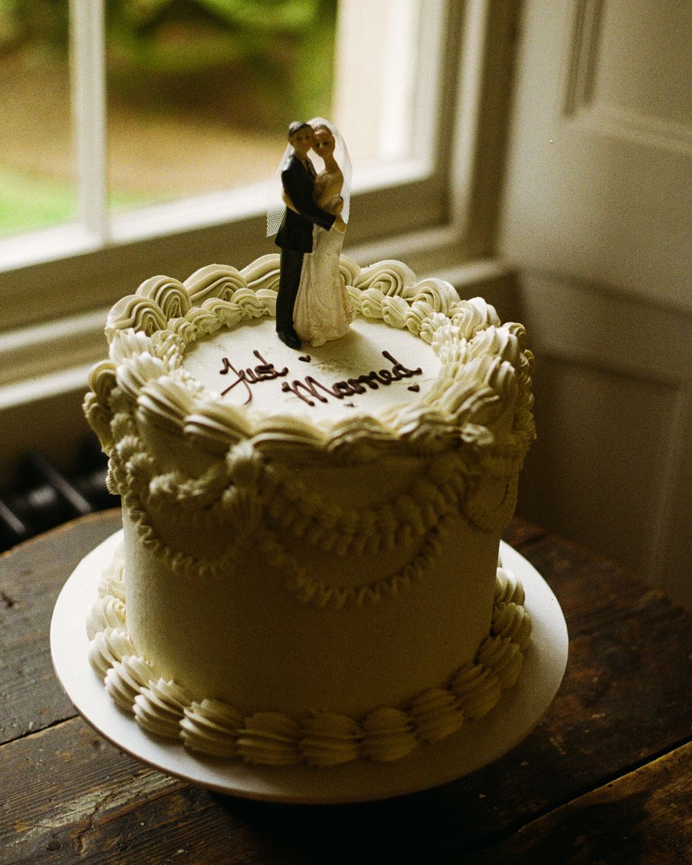 Four sequential photos showing a white wedding cake with delicate piped frosting details on a plate near a window.