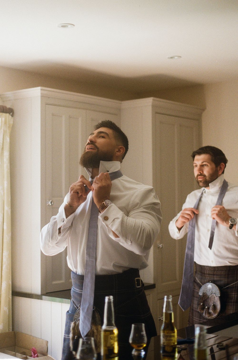 Two groomsmen adjusting their bow ties while getting ready, with beer bottles visible on table in a warm-tinted room.