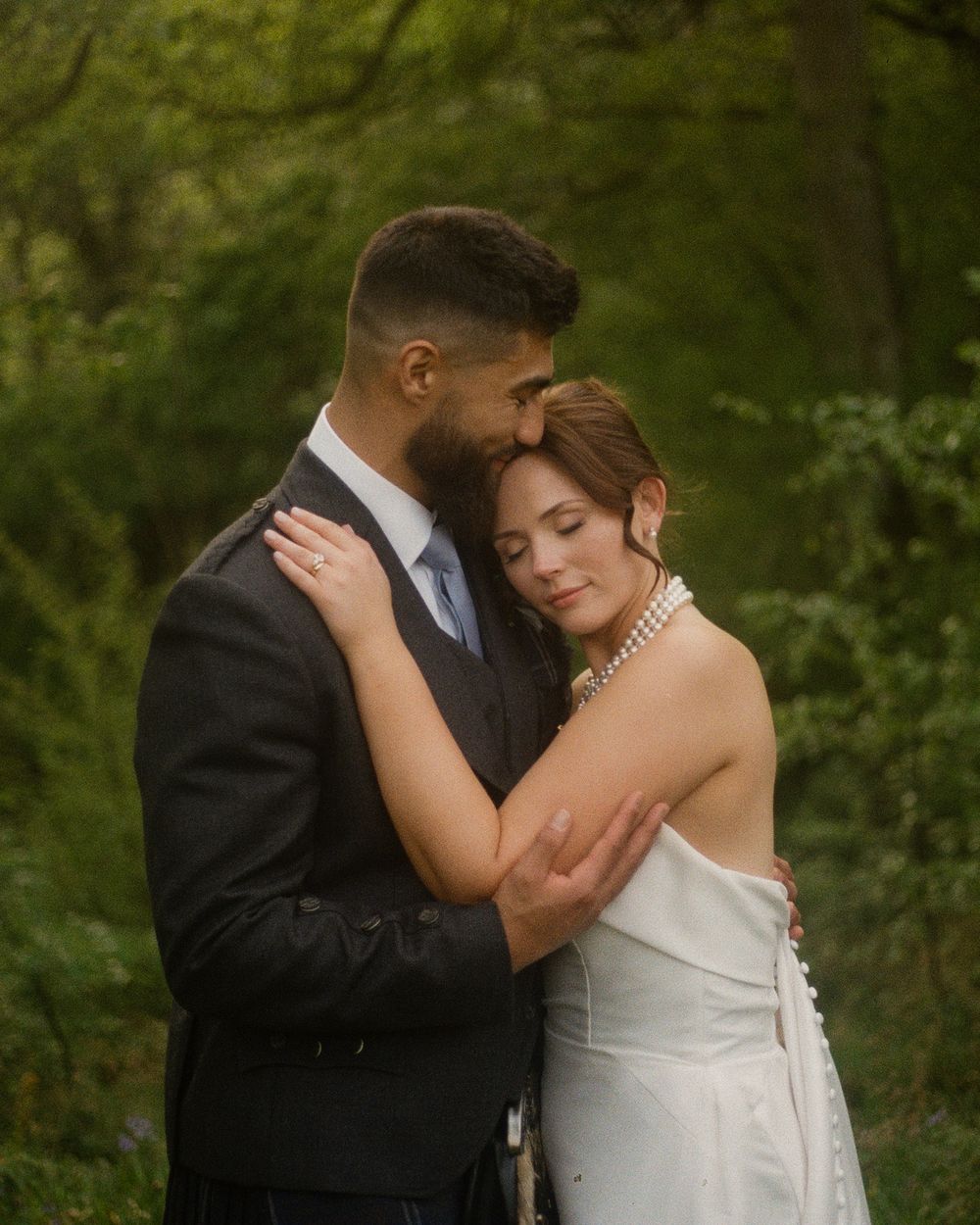 Couple in formal attire sharing an intimate embrace outdoors with lush greenery in the background.