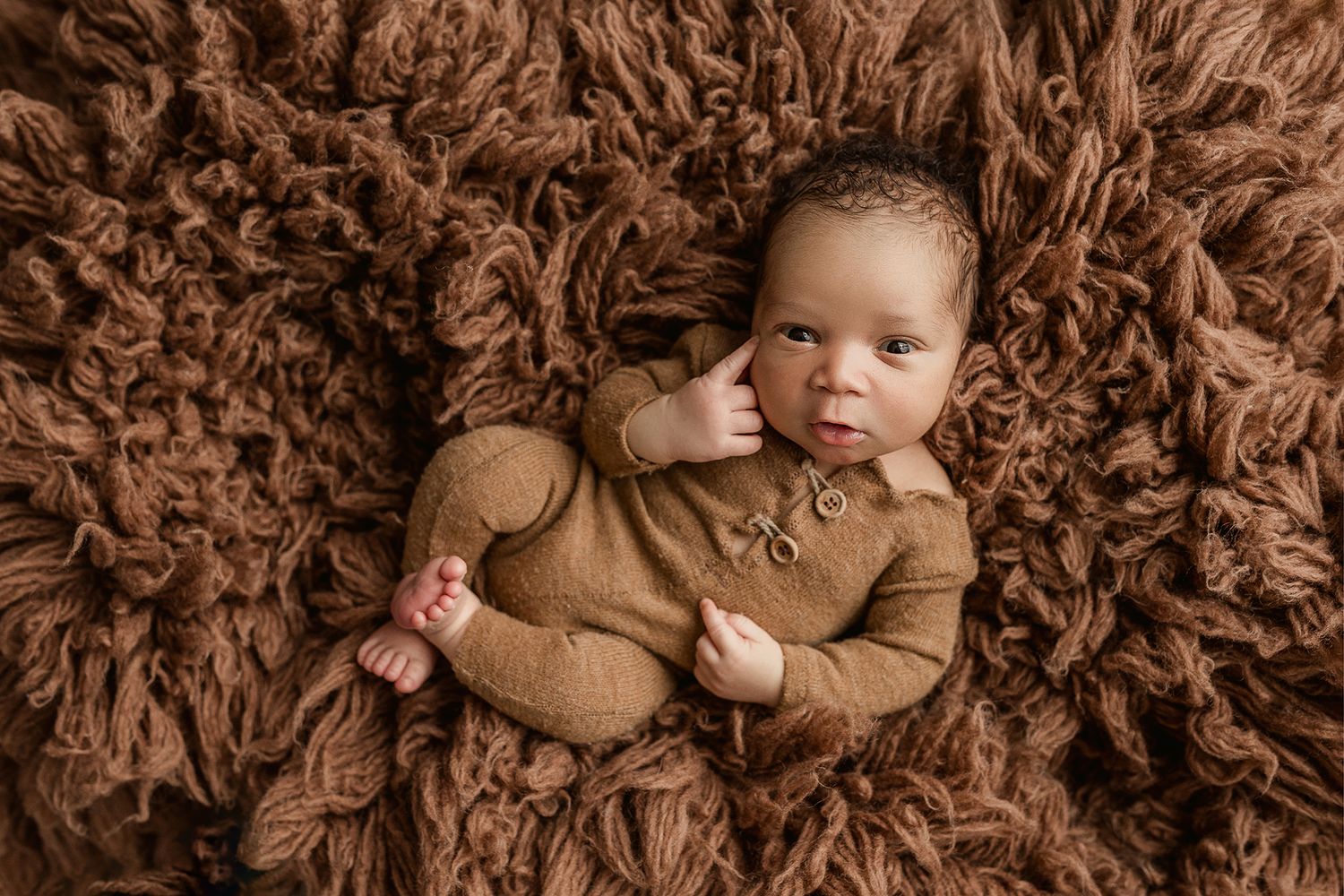 Baby wearing brown outfit lies on textured brown fluffy blanket during professional newborn photo session.