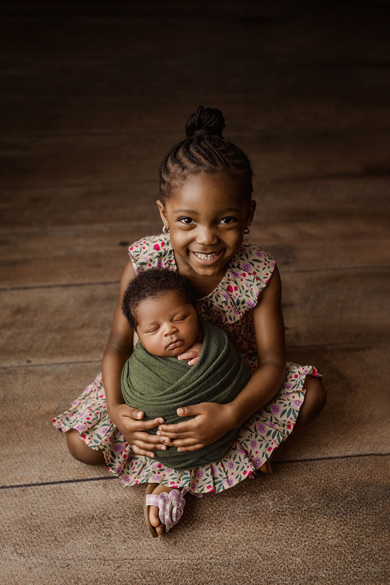 Sweet moment between two siblings as they pose on a wooden floor in a floral dress and olive green swaddle.