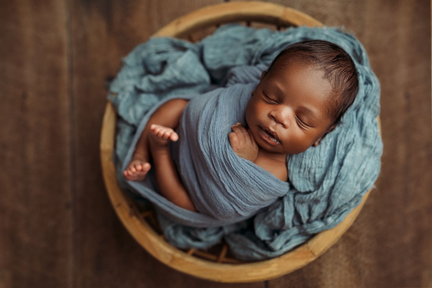 Newborn baby sleeping peacefully in a wooden bowl wrapped in soft turquoise fabric.