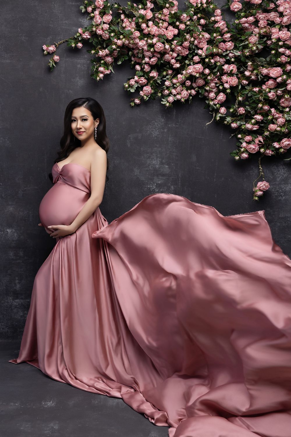 A dusty rose satin maternity gown flows elegantly against a dark wall decorated with pink climbing roses.