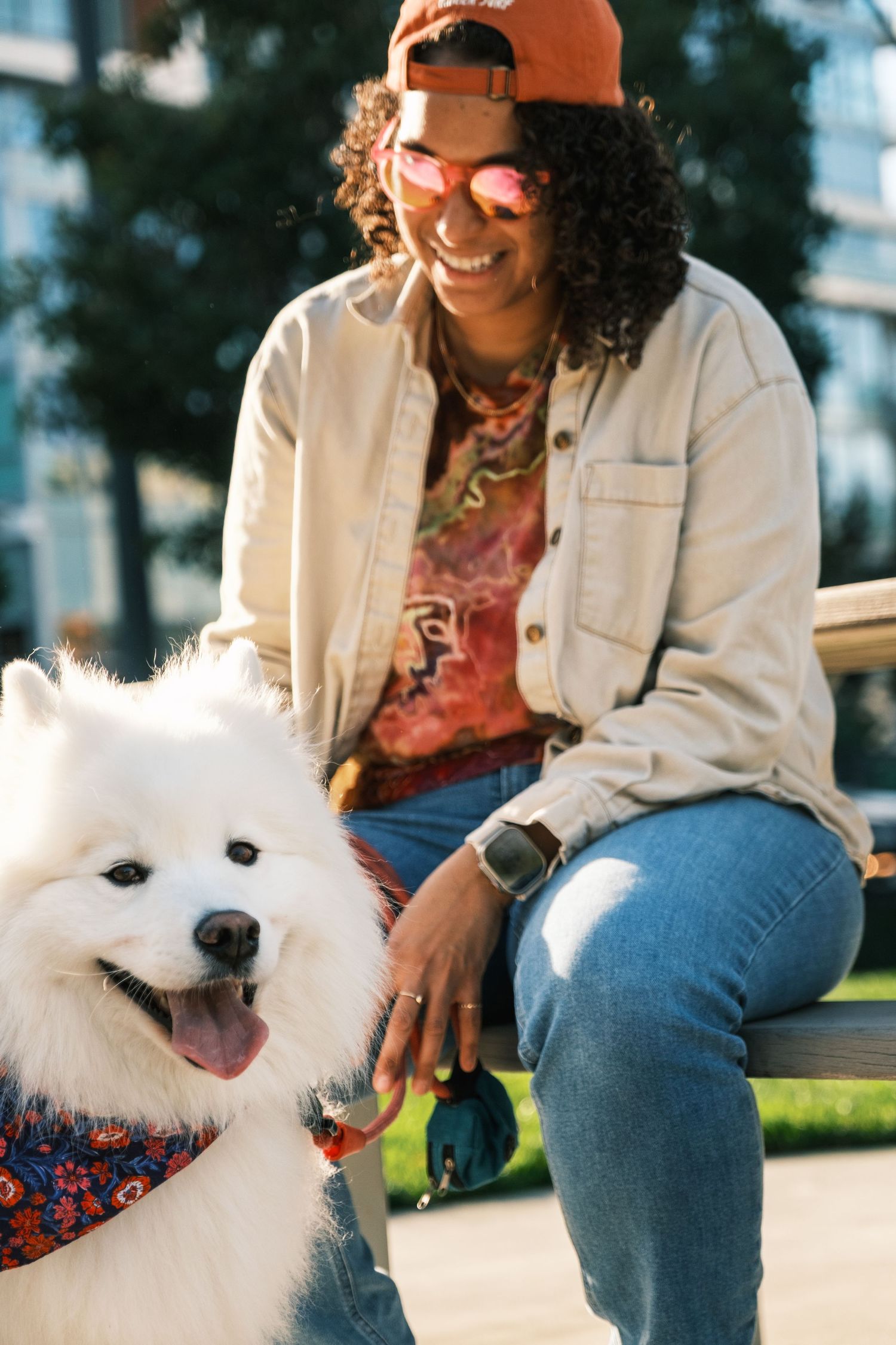 Smiling person in casual denim and sunglasses sits outdoors with happy white Samoyed dog wearing a bandana.
