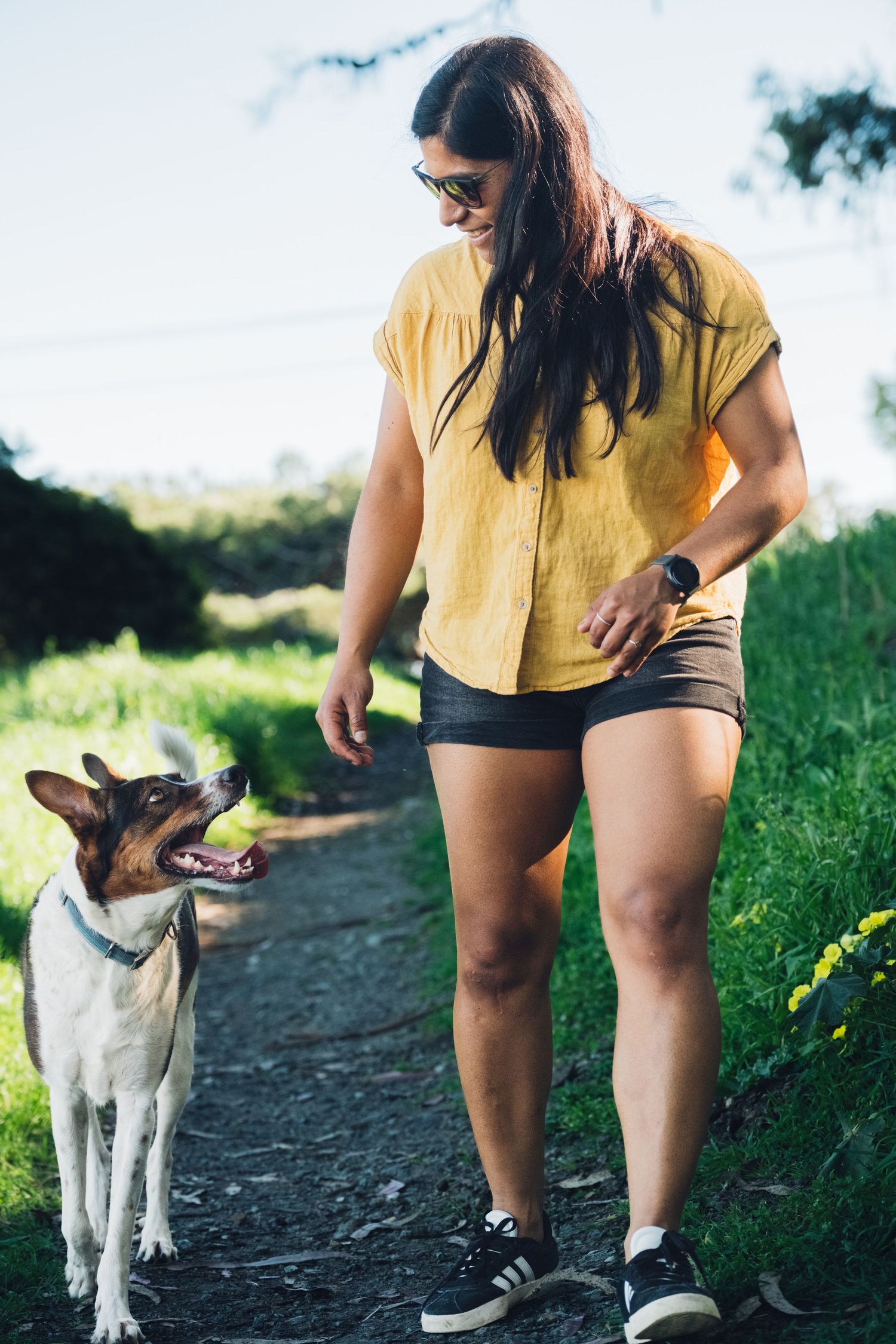 A person in a yellow shirt and shorts stands on a hiking trail with their dog looking upward in a natural setting.