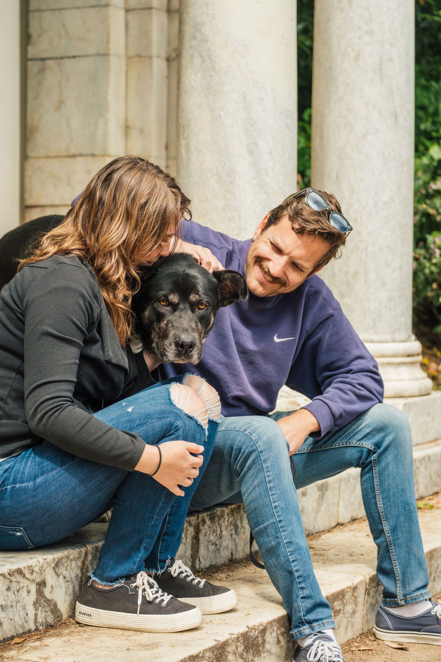A black dog sits between two people on stone steps while they smile and share a tender moment outdoors.