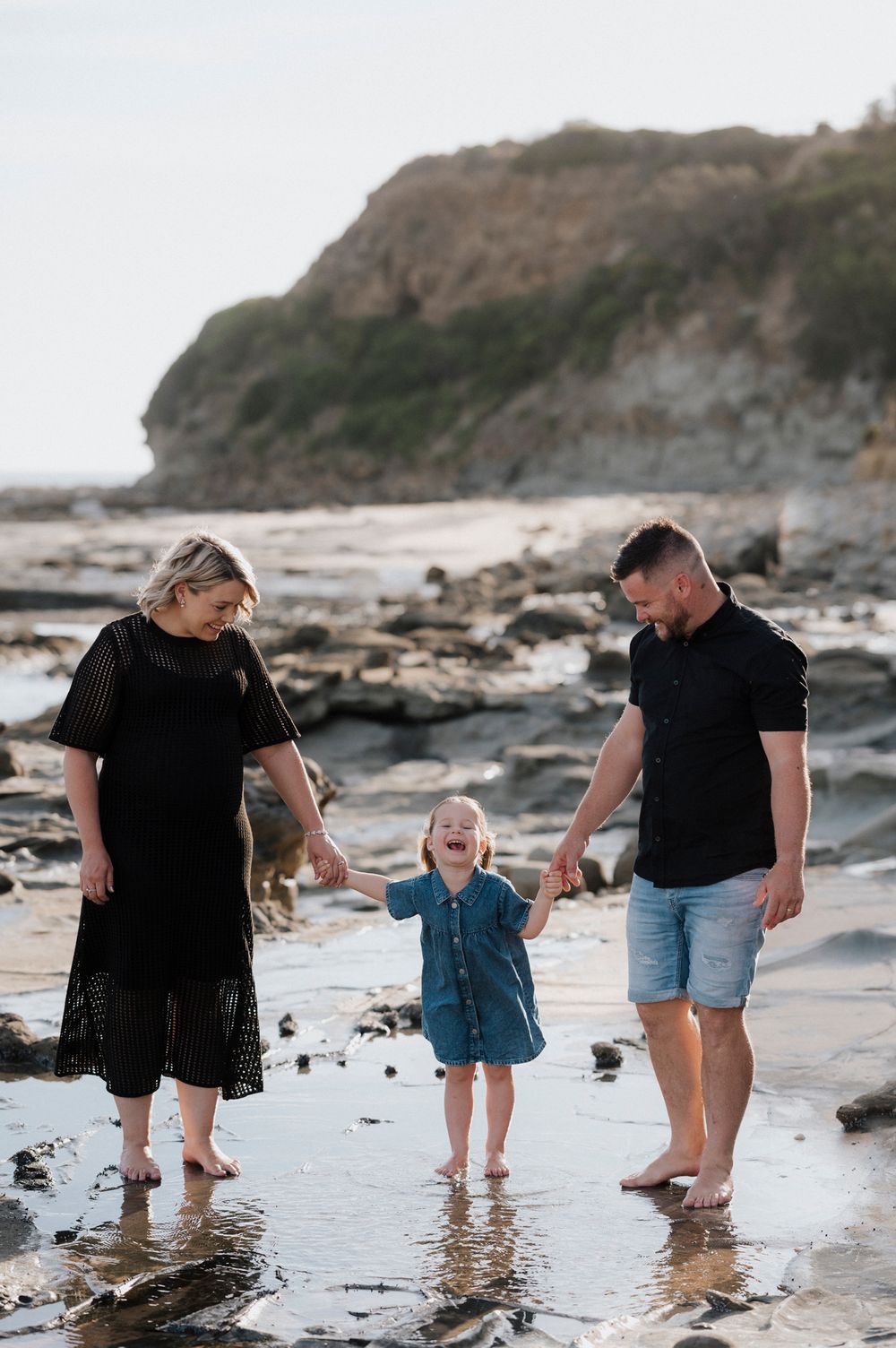A family walks together holding hands along a rocky beach shoreline with cliffs in the background at sunset.
