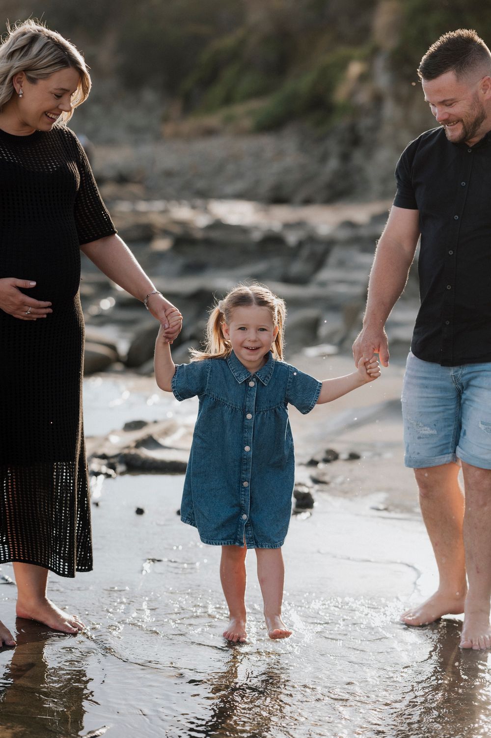A family of three walks together on a rocky beach shoreline at sunset, splashing in shallow water.