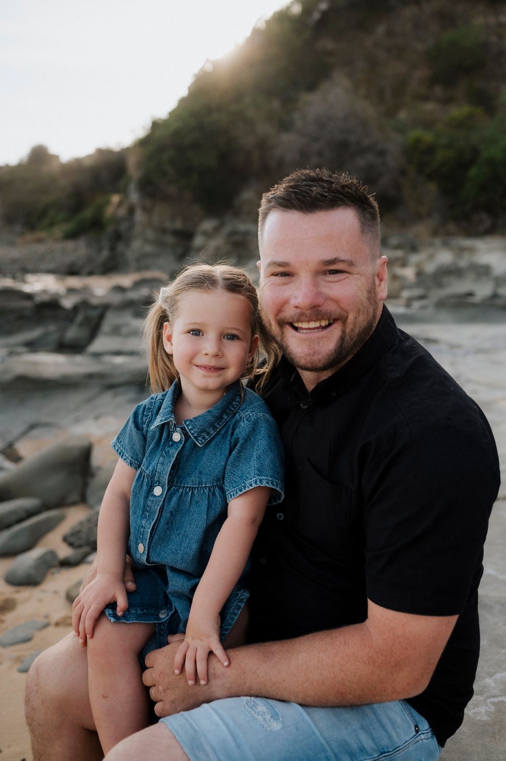 A parent and child sit together on rocky beach at sunset, smiling warmly at the camera.