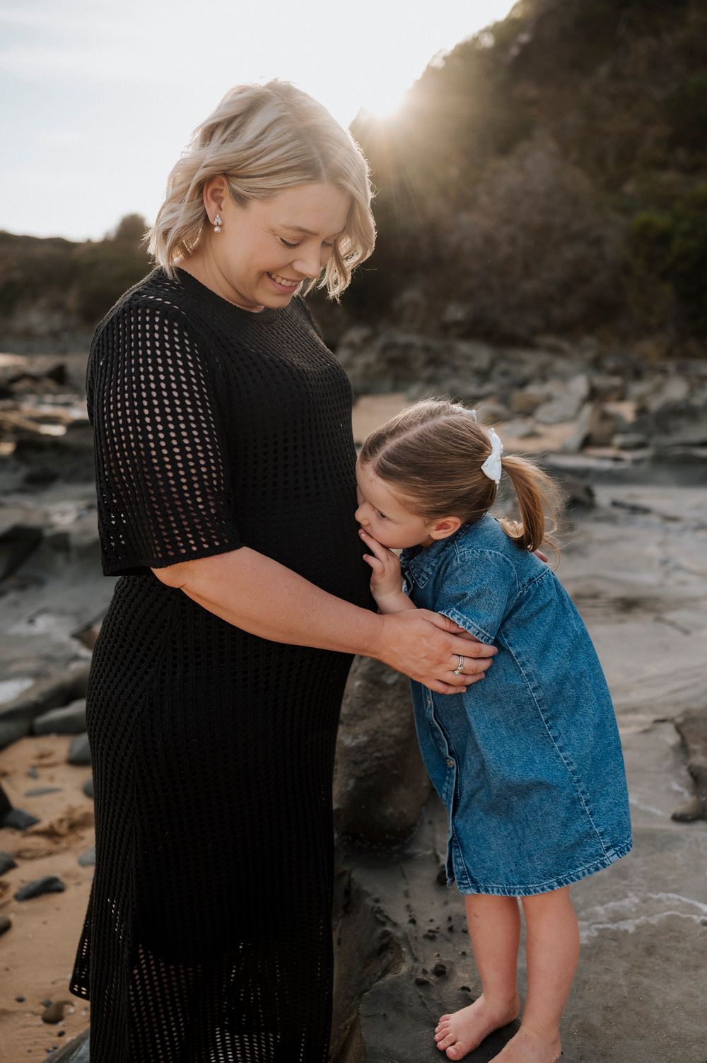 A tender moment between a woman in a black dress and young child in denim on a rocky beach at sunset.