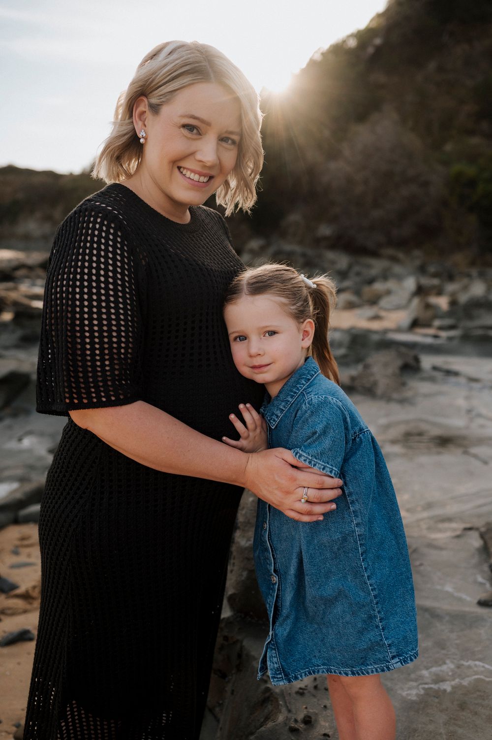 A parent and child share a sweet embrace on a rocky beach at sunset in a heartwarming outdoor portrait.
