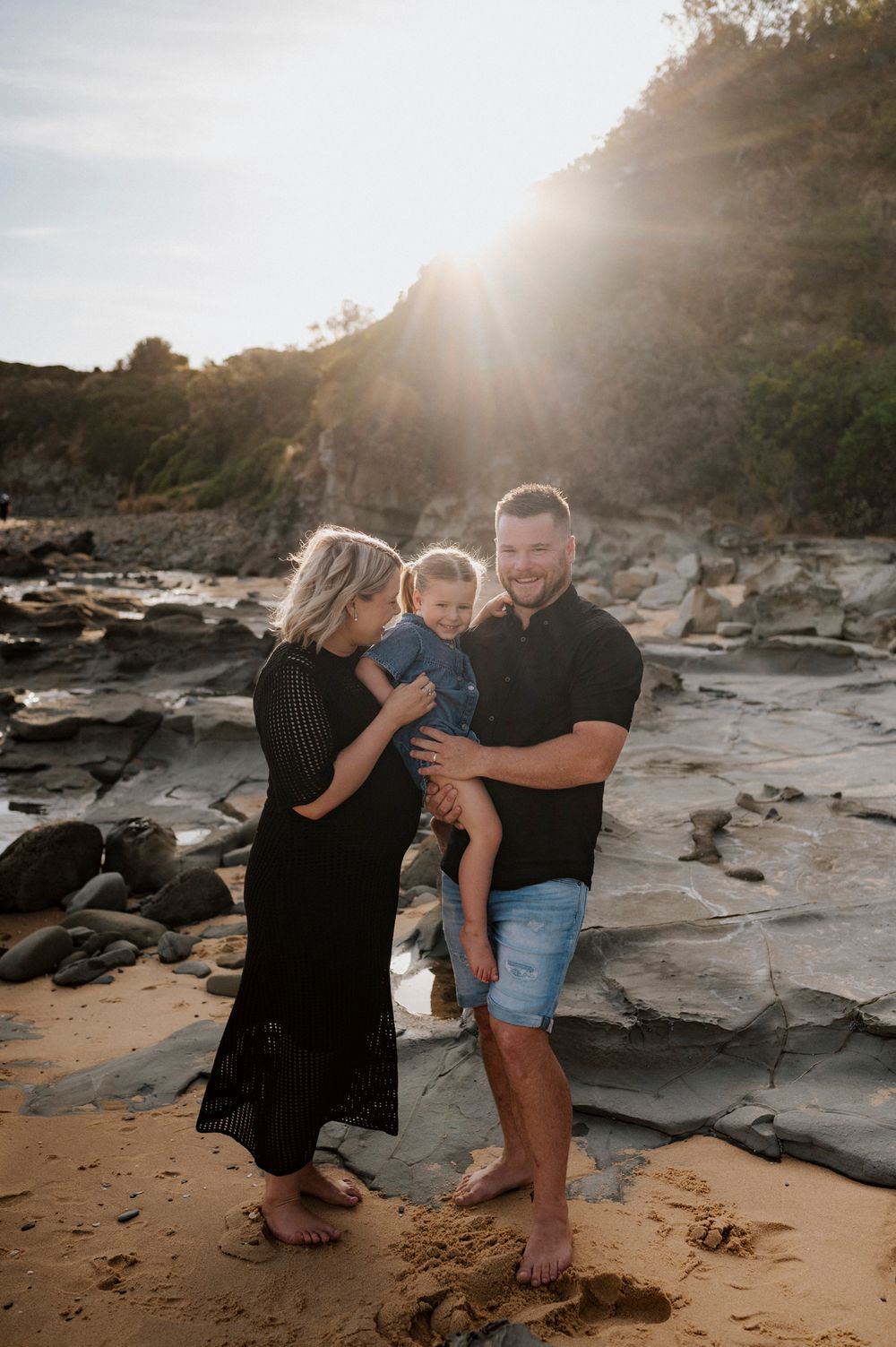 Family enjoying sunset together on a rocky beach with sunlight streaming through cliffs in the background.