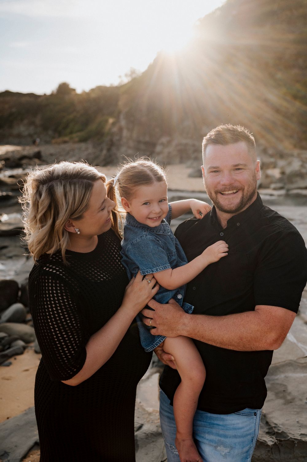 A family enjoys a sunset moment together on a rocky beach with sunlight streaming through the background mountains.
