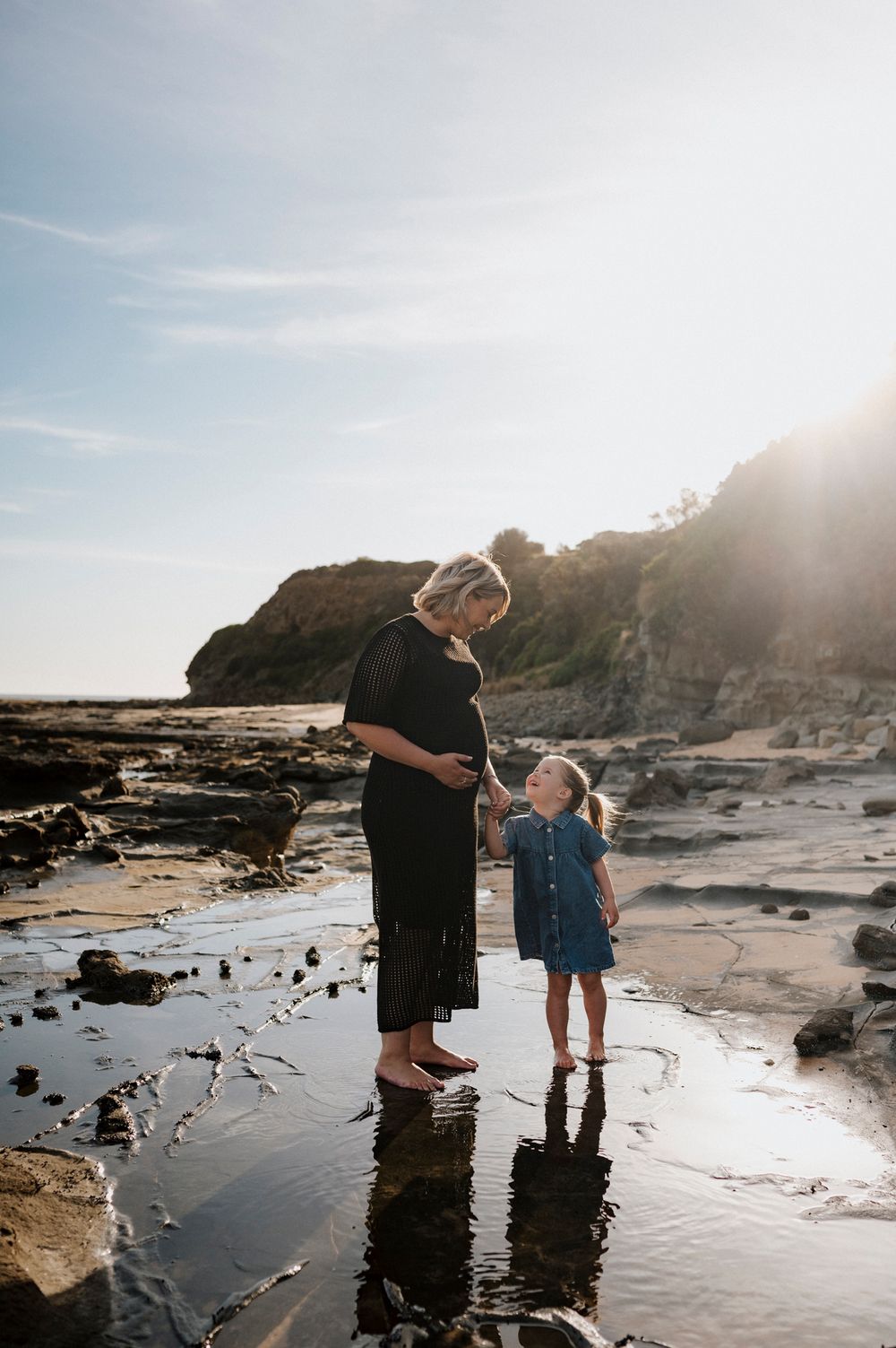 Two figures stand on rocky beach tide pools at sunset with coastal cliffs in background, light glowing through clouds.