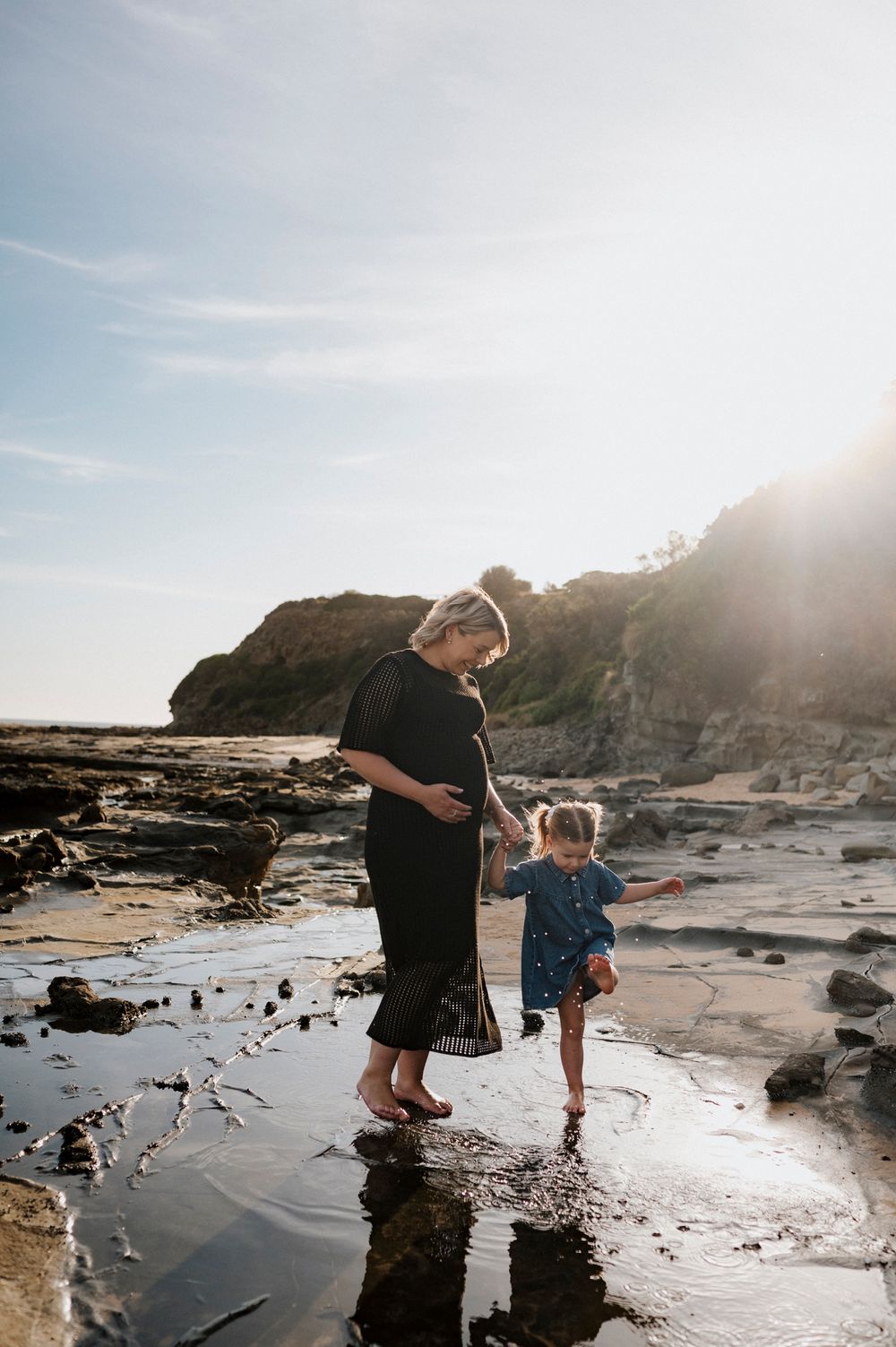 Two people explore tide pools together on a rocky beach at sunset with cliffs and ocean in the background.