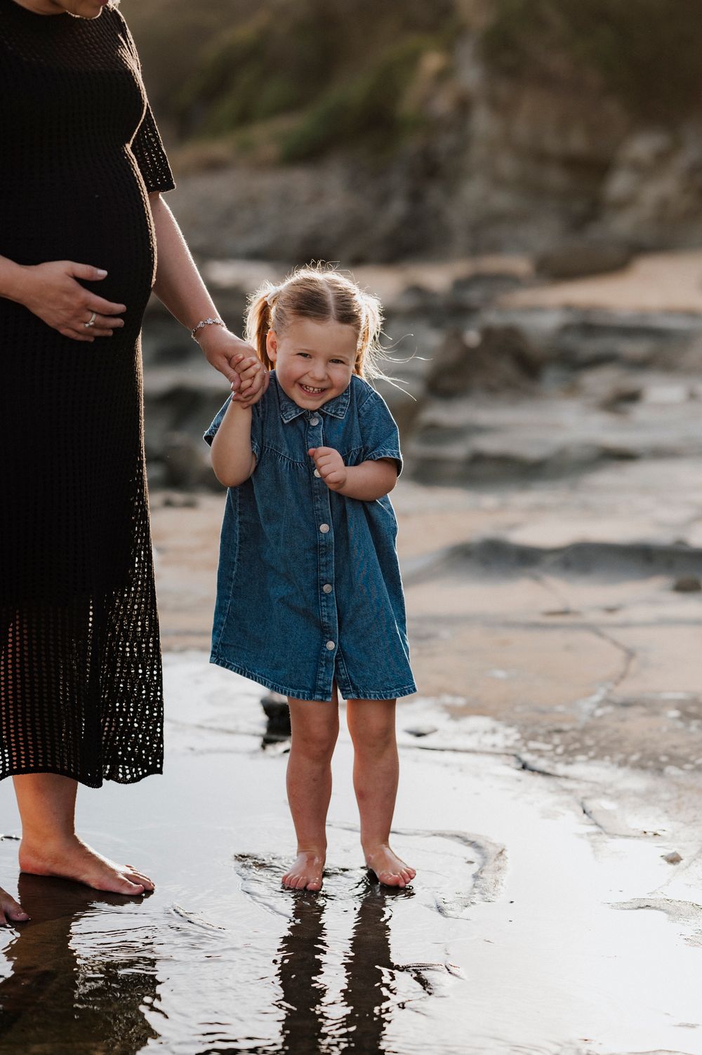 A young child in a denim dress splashes in shallow water at the beach while holding an adult's hand at sunset.