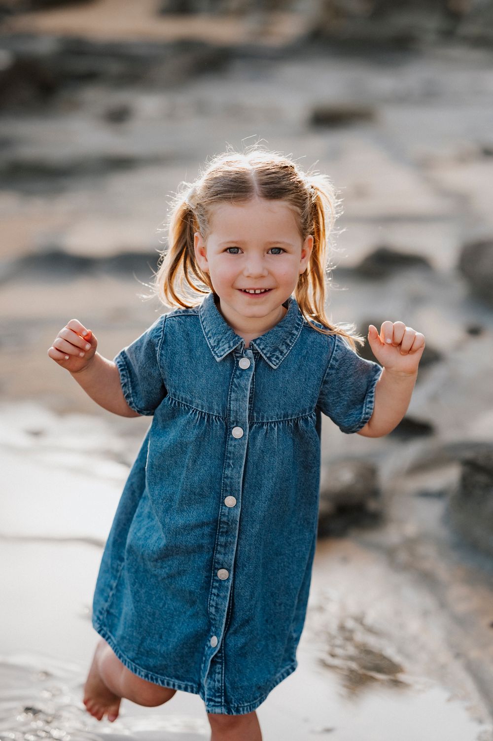 A toddler wearing a denim dress plays joyfully on a rocky beach at sunset.