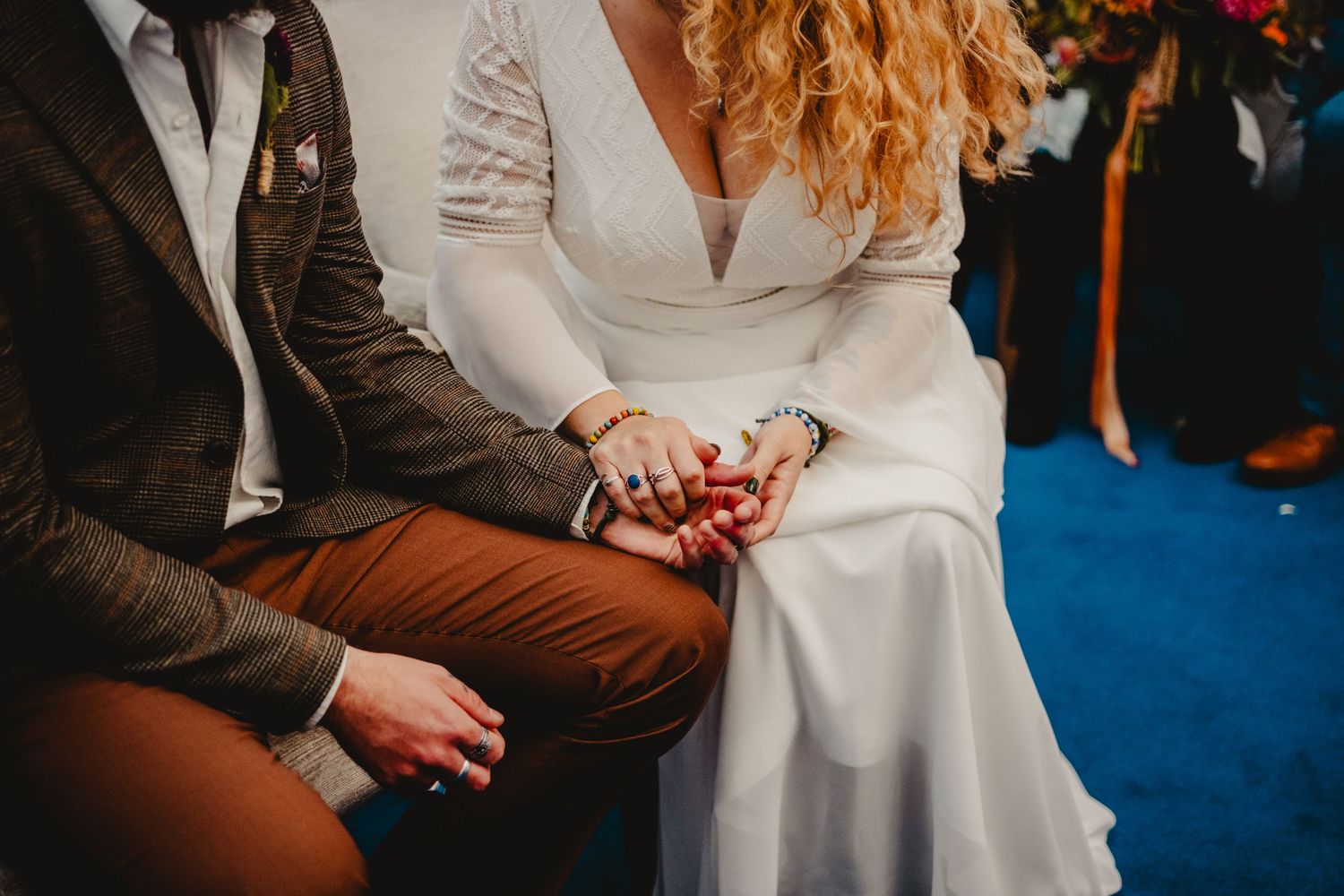 Close-up shot of couple holding hands at wedding ceremony with bride wearing elegant white dress and groom in brown suit.
