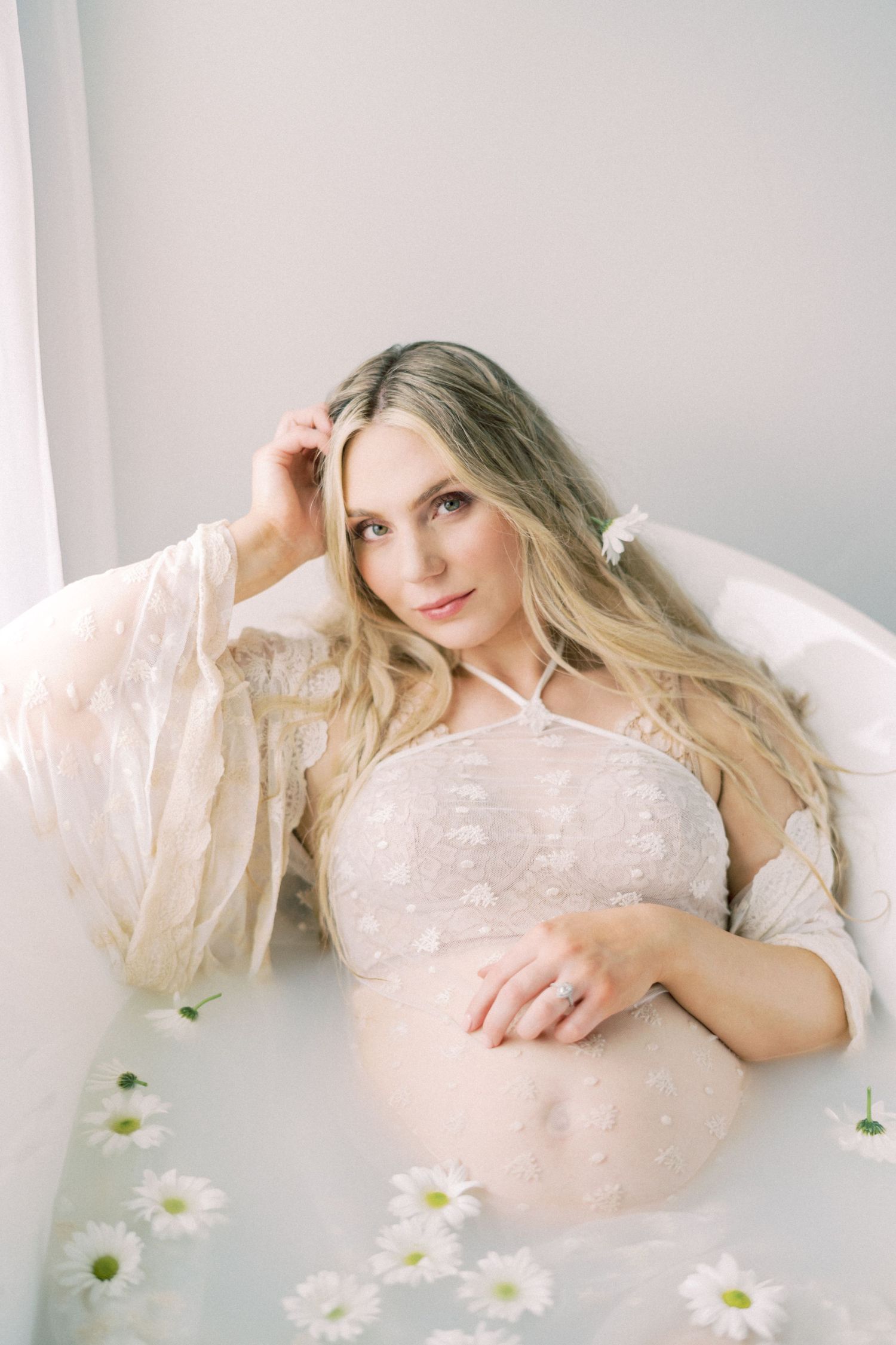 Ethereal maternity photo with flowing sheer robe and scattered daisies in dreamy soft lighting.