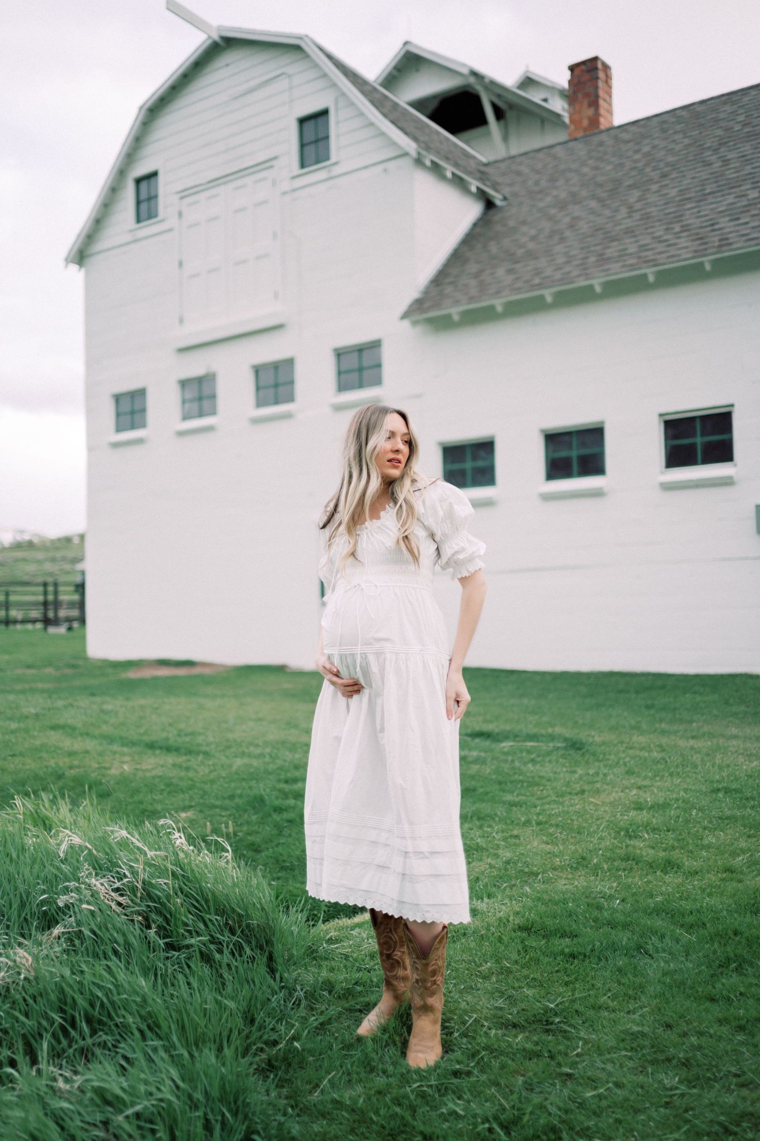 Someone in a flowing white dress stands barefoot in a green mountain meadow on an overcast day.