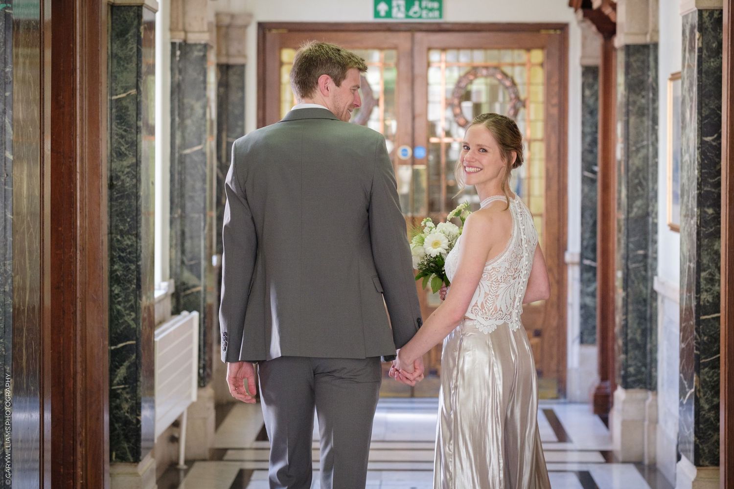 A couple in wedding attire holds hands while walking down an elegant hallway with marble columns.