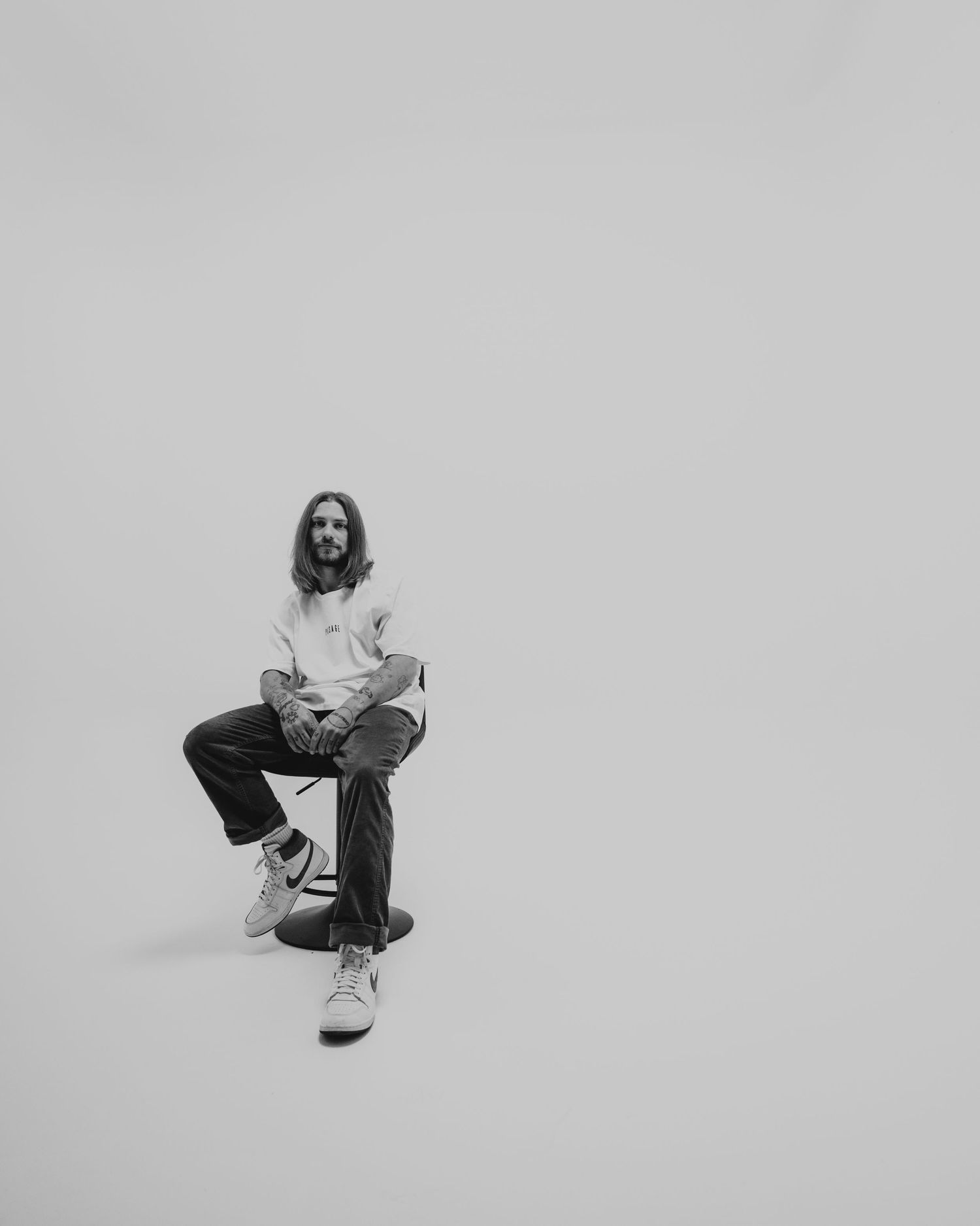 Black and white minimalist photo of a person sitting on a stool against a plain wall wearing casual attire.