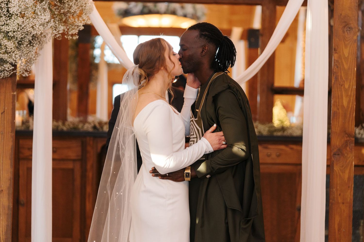 A loving couple shares an intimate moment during their wedding ceremony in a rustic wooden venue decorated with white draping.