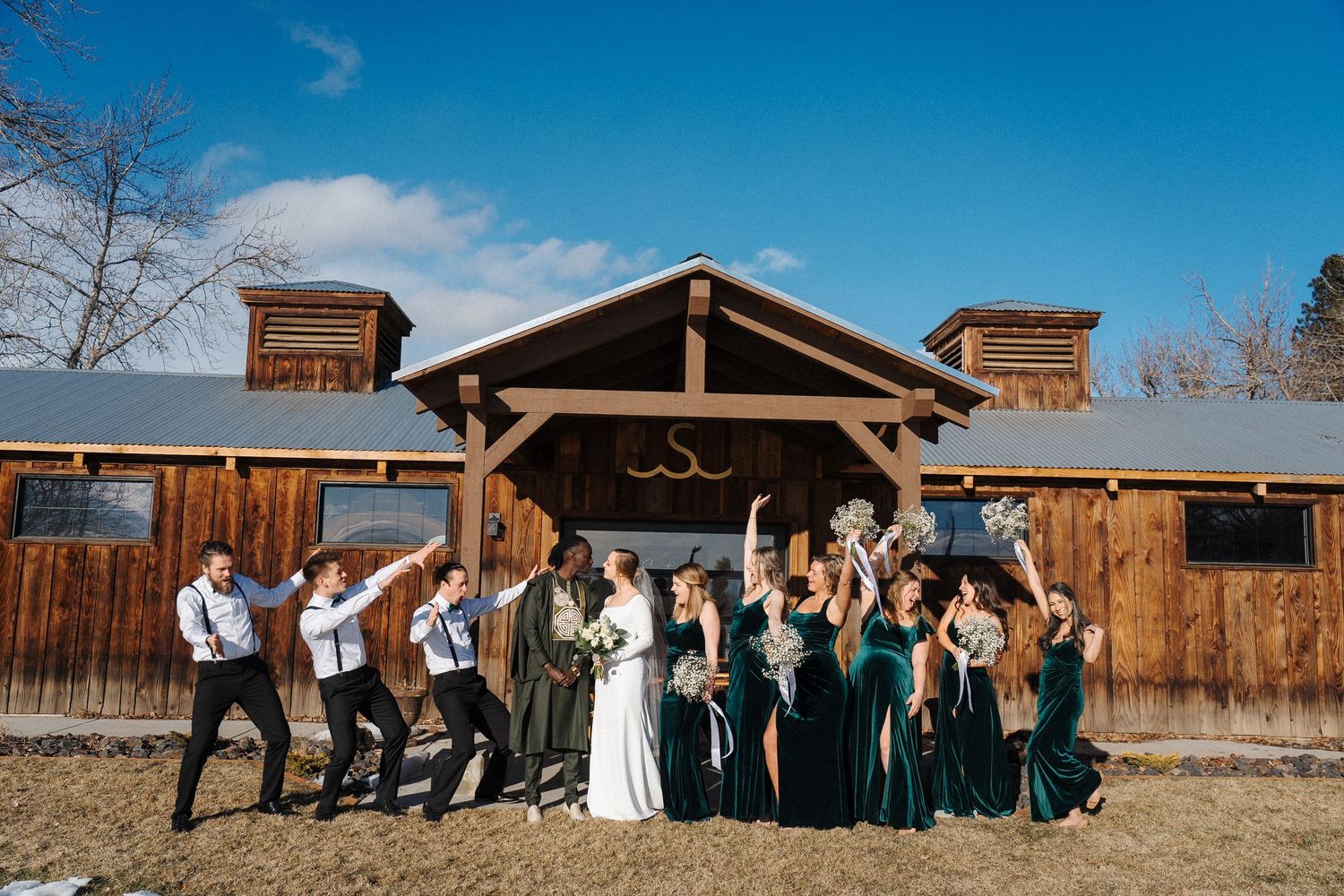 Wedding party celebrates joyfully in front of rustic wooden barn venue under blue sky.