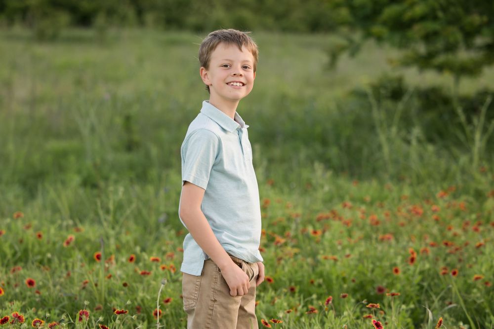 A young person stands in a sunny meadow with wildflowers, wearing a light blue shirt and tan pants on a summer day.