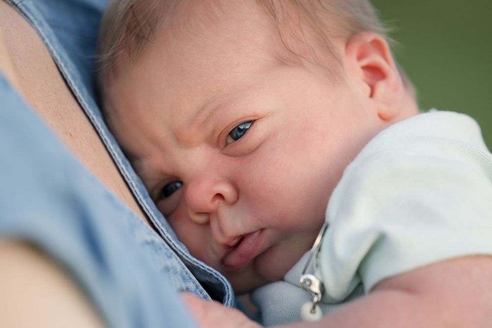 Close-up view of a young baby in a white onesie resting against someone wearing denim.