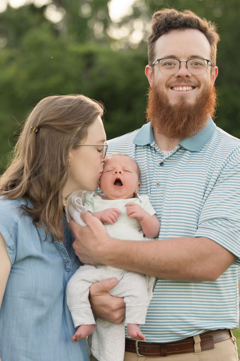 A family portrait outdoors showing a couple holding a yawning baby in white clothes against a green background.