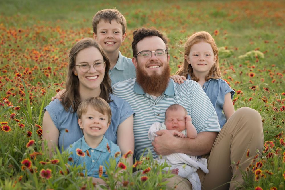 A family group smiles while sitting together in a field of red Indian Paintbrush flowers.
