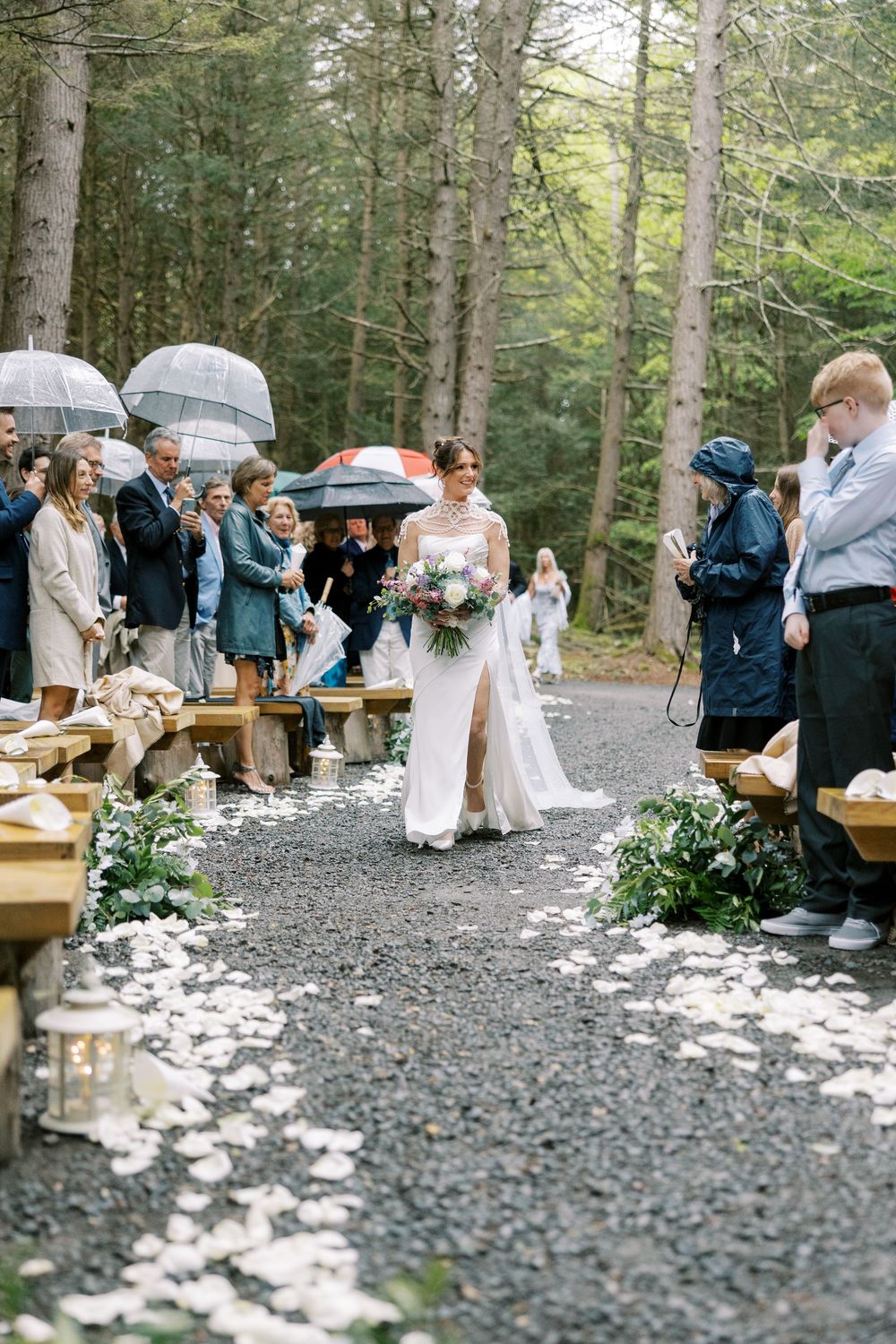 A rainy outdoor wedding ceremony in a forest with guests holding umbrellas along a path lined with flowers and benches at Windham Manor.