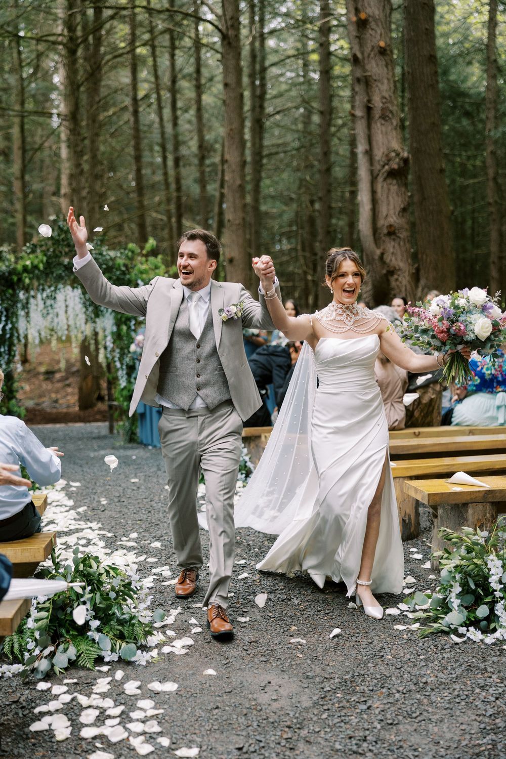 A joyful couple celebrates while walking down a forest path decorated with white petals and floral arrangements at Windham Manor.