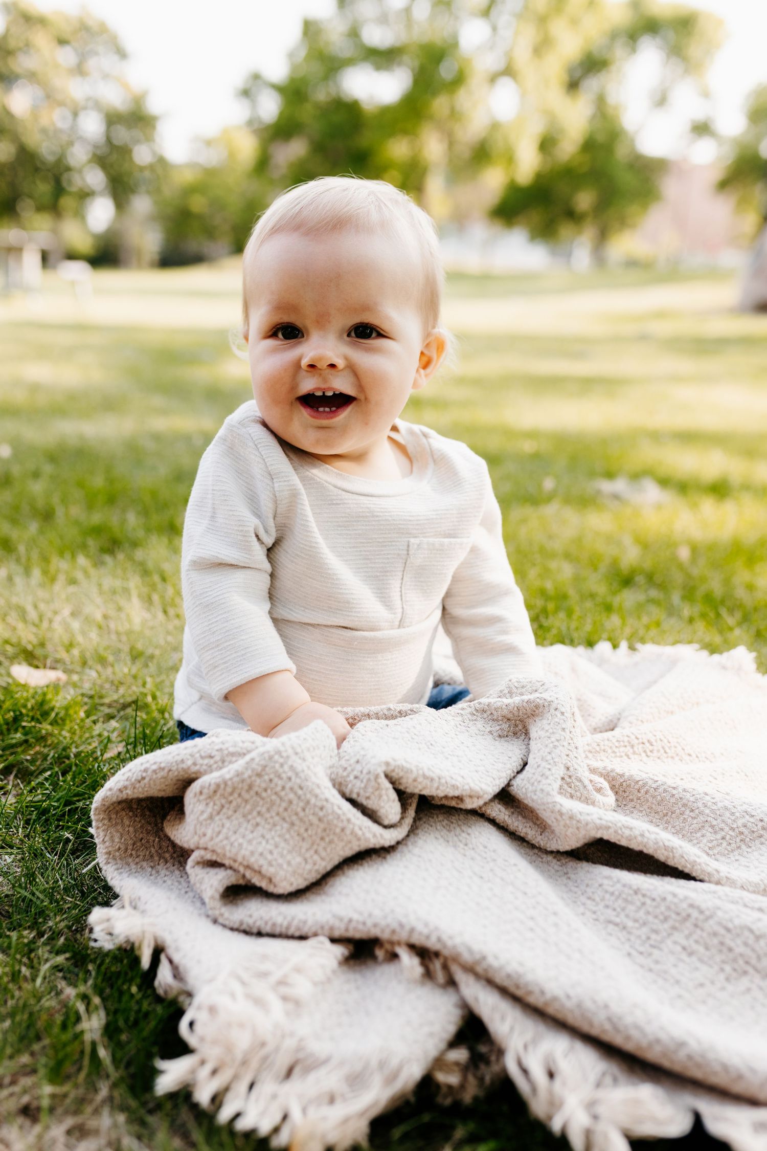 A baby wearing a cream-colored sweater sits on a knit blanket on grass, smiling joyfully outdoors on a sunny day.