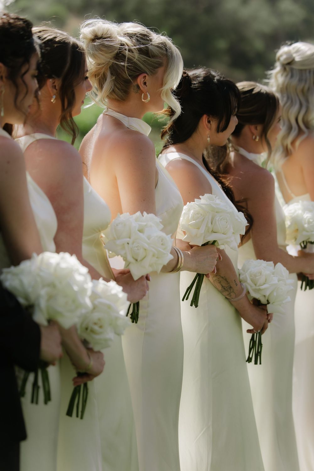 Row of bridesmaids in elegant white dresses holding beautiful white peony bouquets at an outdoor wedding ceremony.