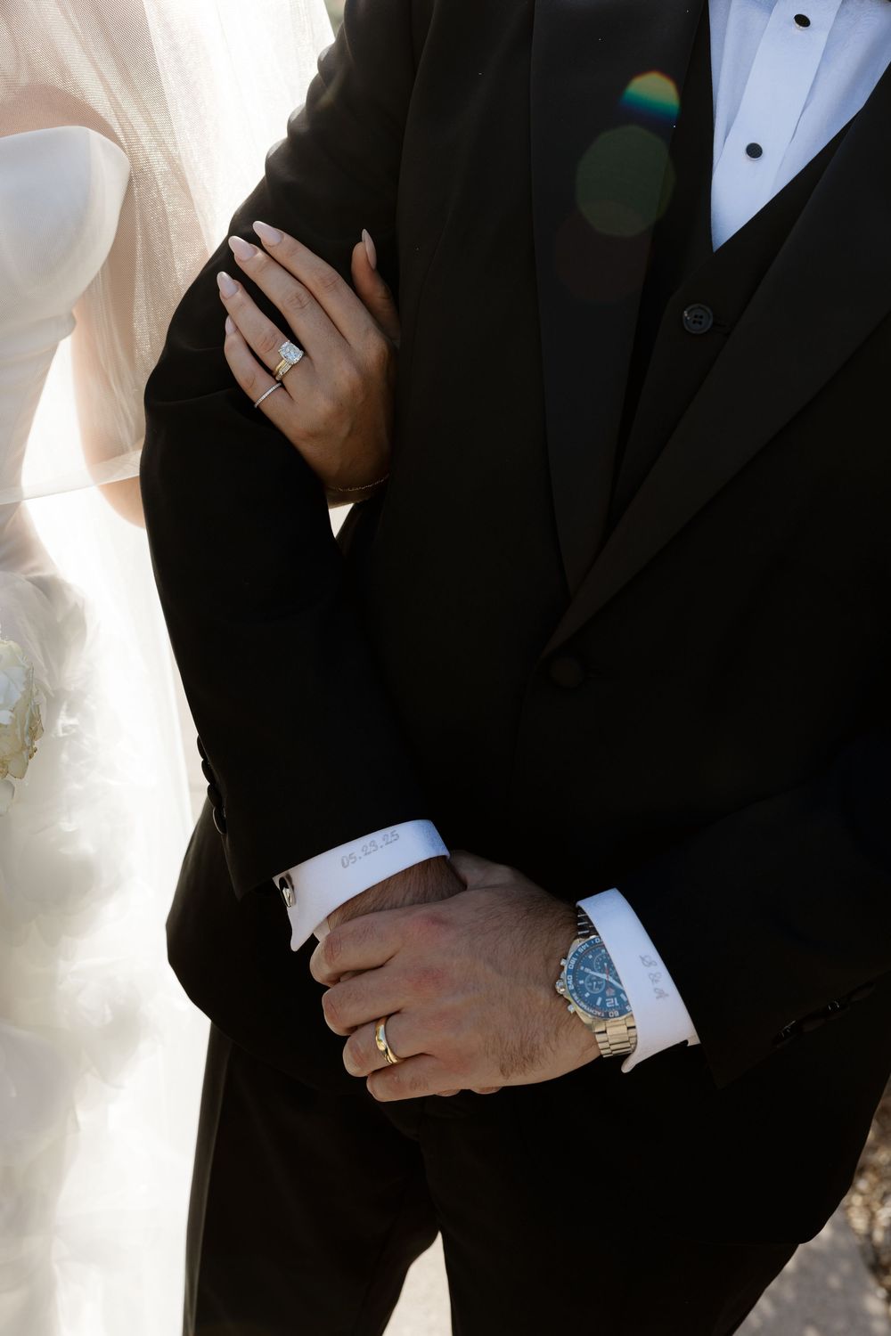 Close-up view of bride and groom's hands showing wedding rings and elegant watch against black tuxedo.