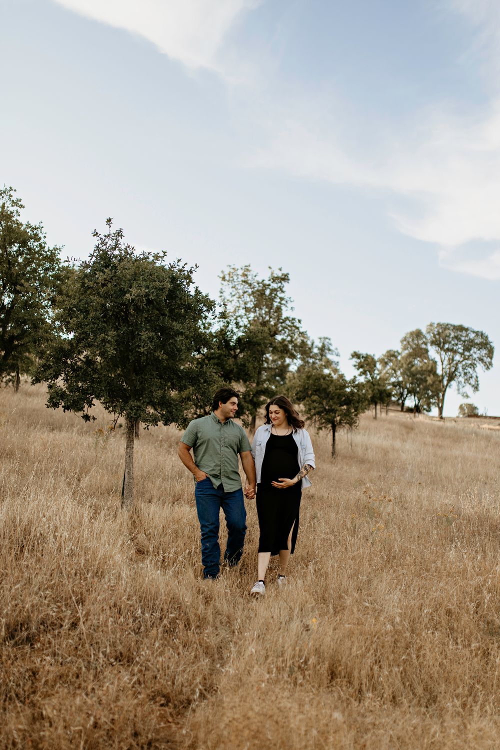 Couple portraits walking together through a golden dry grass field with scattered oak trees under a bright sky in EDH
