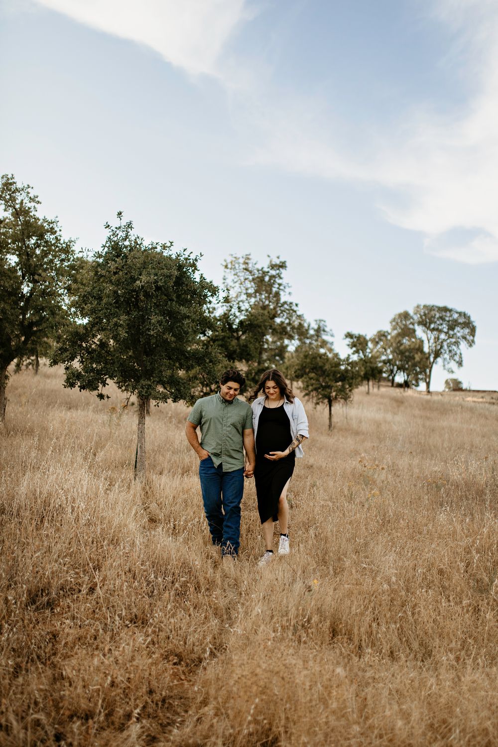 A couple stands together in a golden field with scattered oak trees on a sunny afternoon against a blue sky for photoshoot.