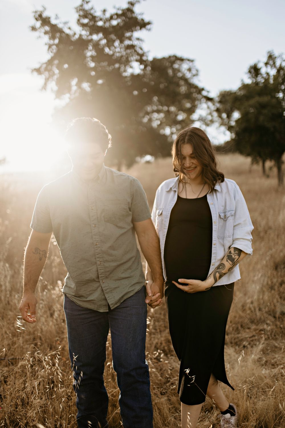 A couple holds hands while walking through a sun-drenched golden field at sunset for maternity pictures in Cameron Park.