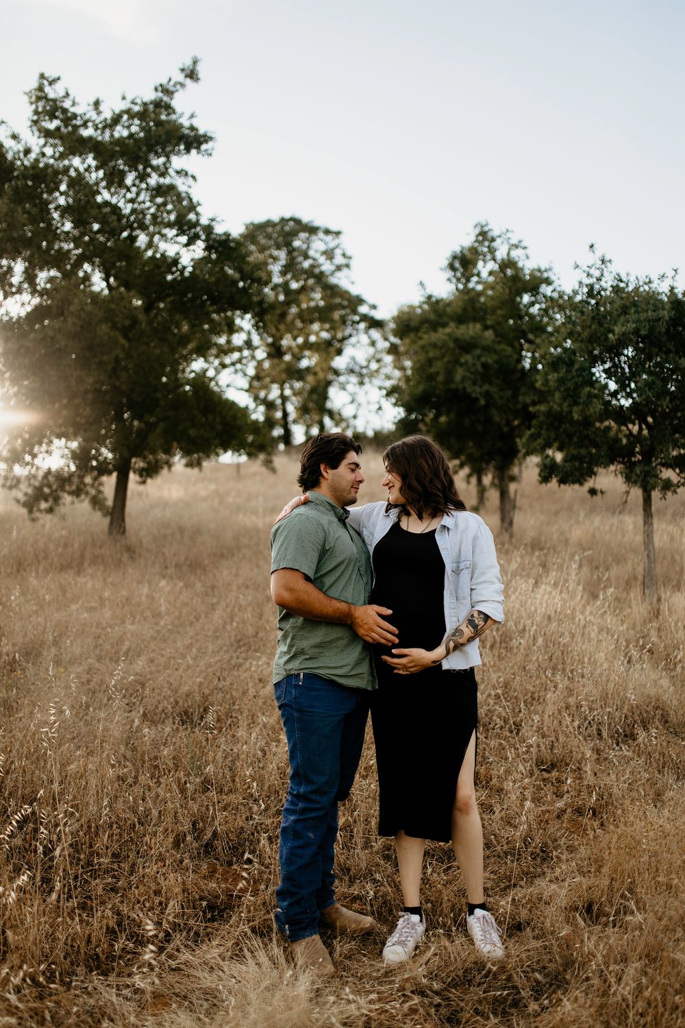 A couple shares an intimate moment in a golden field at sunset with trees silhouetted in the background for maternity photos.