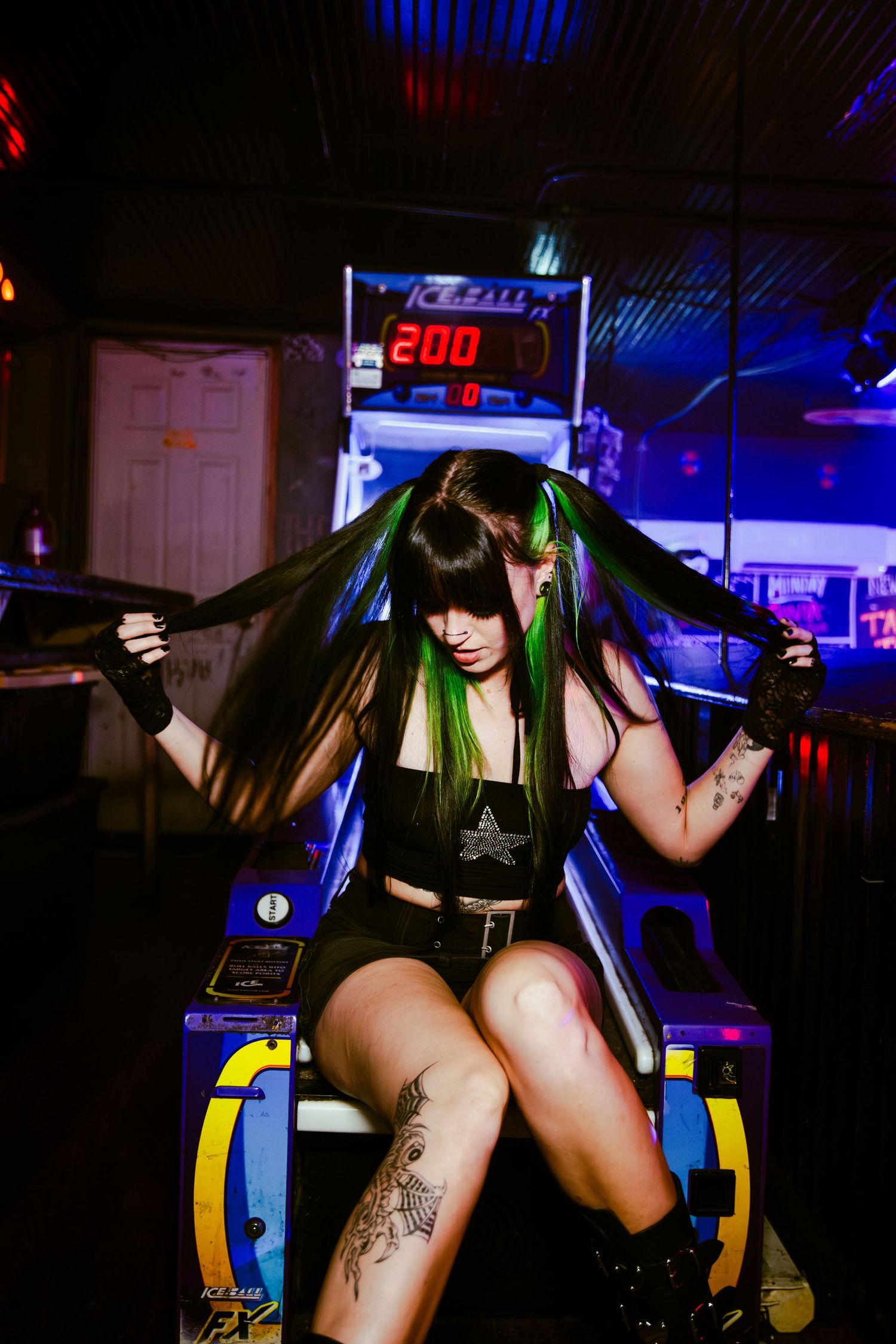 A person in dark clothing with green hair streaks poses on an arcade game machine in a dimly lit venue with neon lights.
