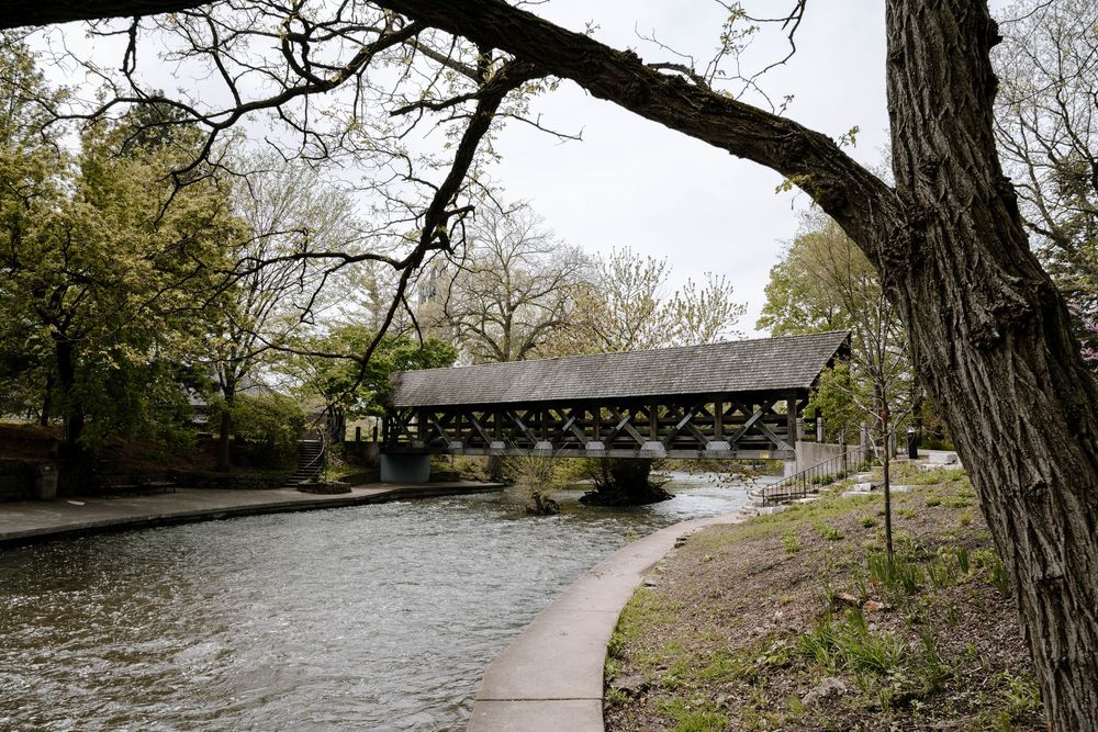 A rustic wooden covered bridge spans over a winding river path with trees and foliage in the background.