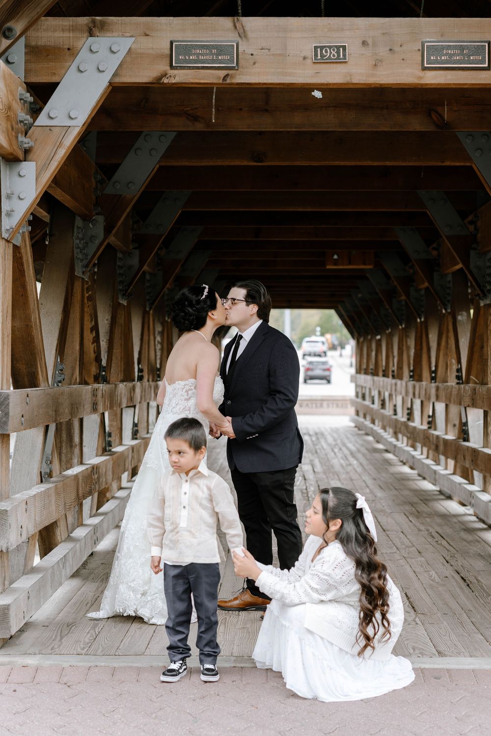 A wedding photo of a family embracing inside a wooden covered bridge with rustic beams.