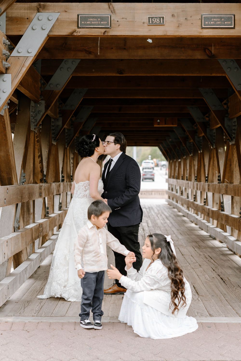 Wedding couple and children share a tender moment inside a covered wooden bridge.