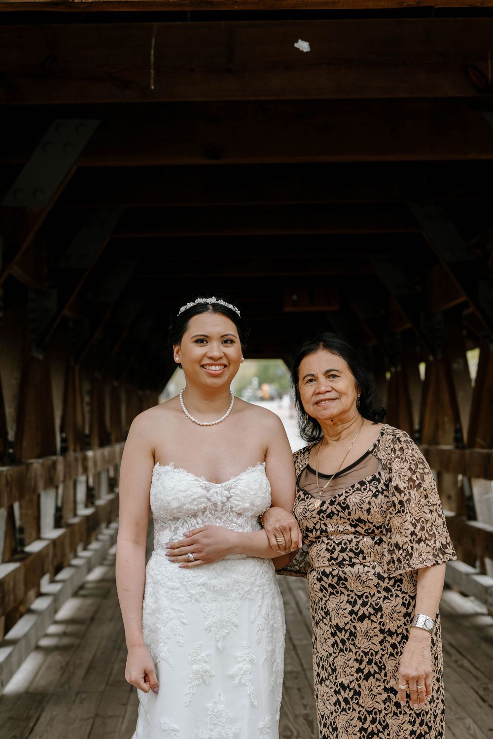 A bride in white dress and veil poses with a woman in a floral dress inside a covered bridge.