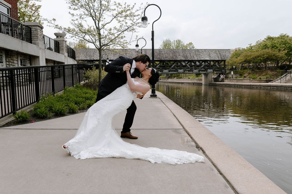 A romantic wedding photo captured on a waterfront path with a covered bridge in the background.
