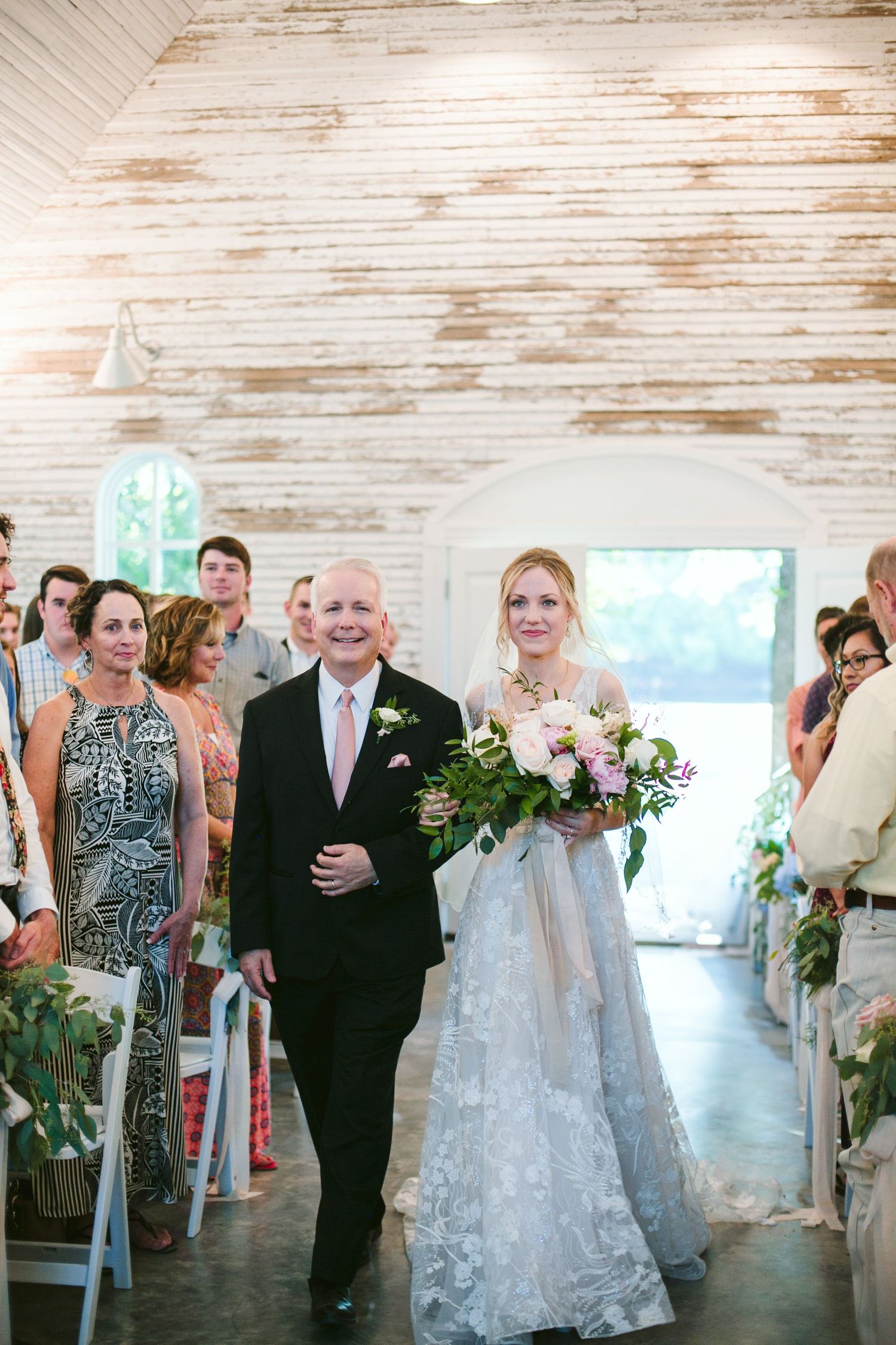 A procession walks down the aisle at a rustic barn venue with whitewashed wooden walls and natural light.